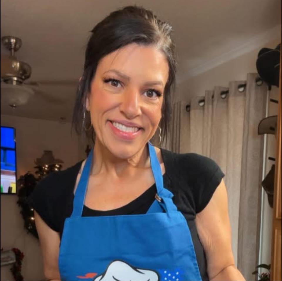 A woman with dark hair in a ponytail smiles at the camera, wearing a black shirt and a blue apron, standing indoors with neutral-colored curtains and a ceiling fan in the background.