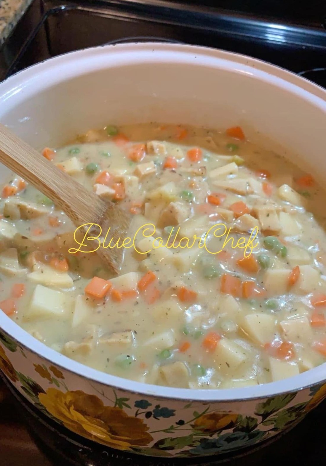 A pot of creamy soup with diced potatoes, carrots, peas, and chicken is being stirred with a wooden spoon. The pot has a floral pattern on its side.