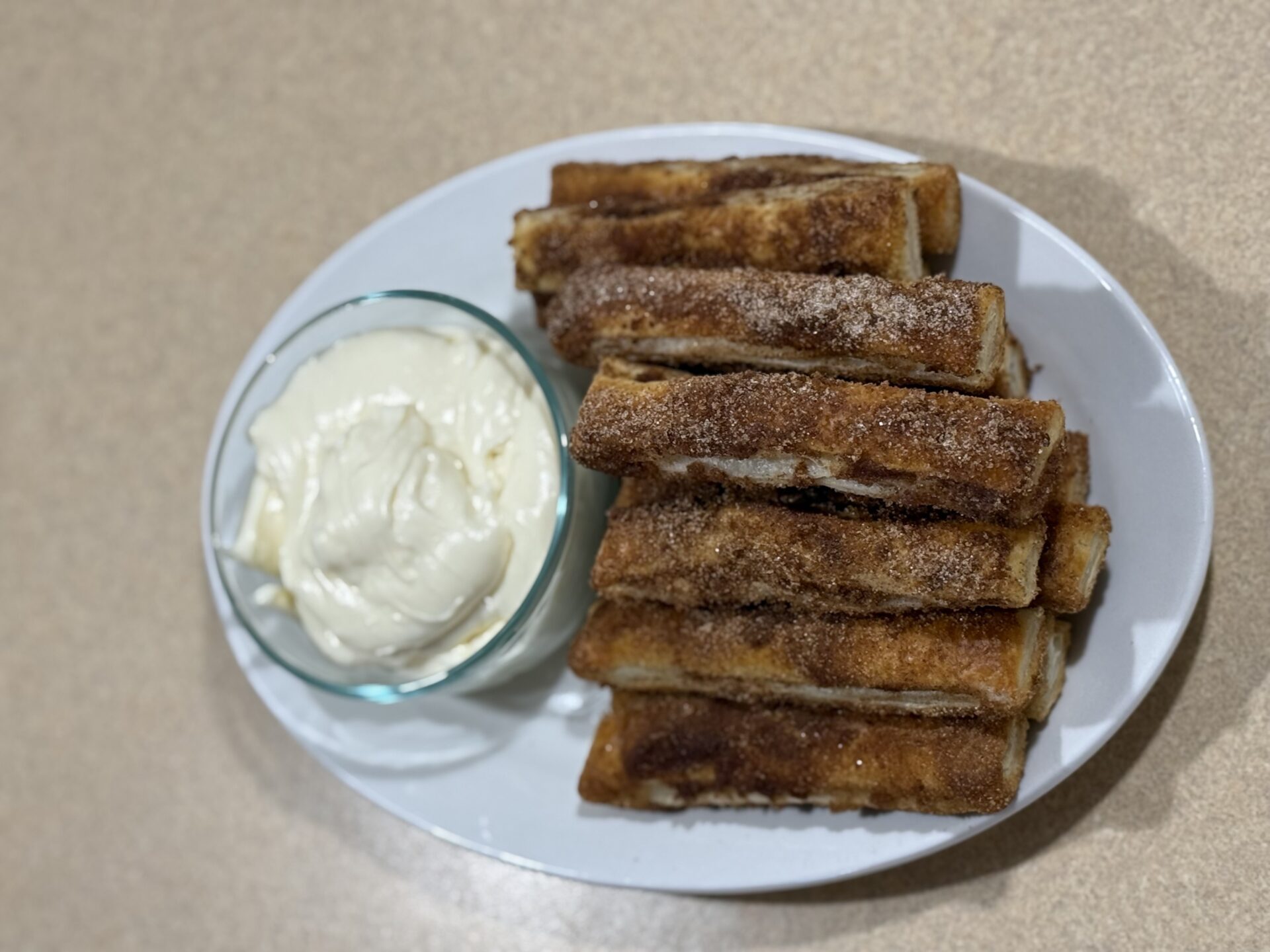 A white oval plate with cinnamon sugar-coated toast sticks beside a small glass bowl of creamy white dip, all placed on a beige countertop.