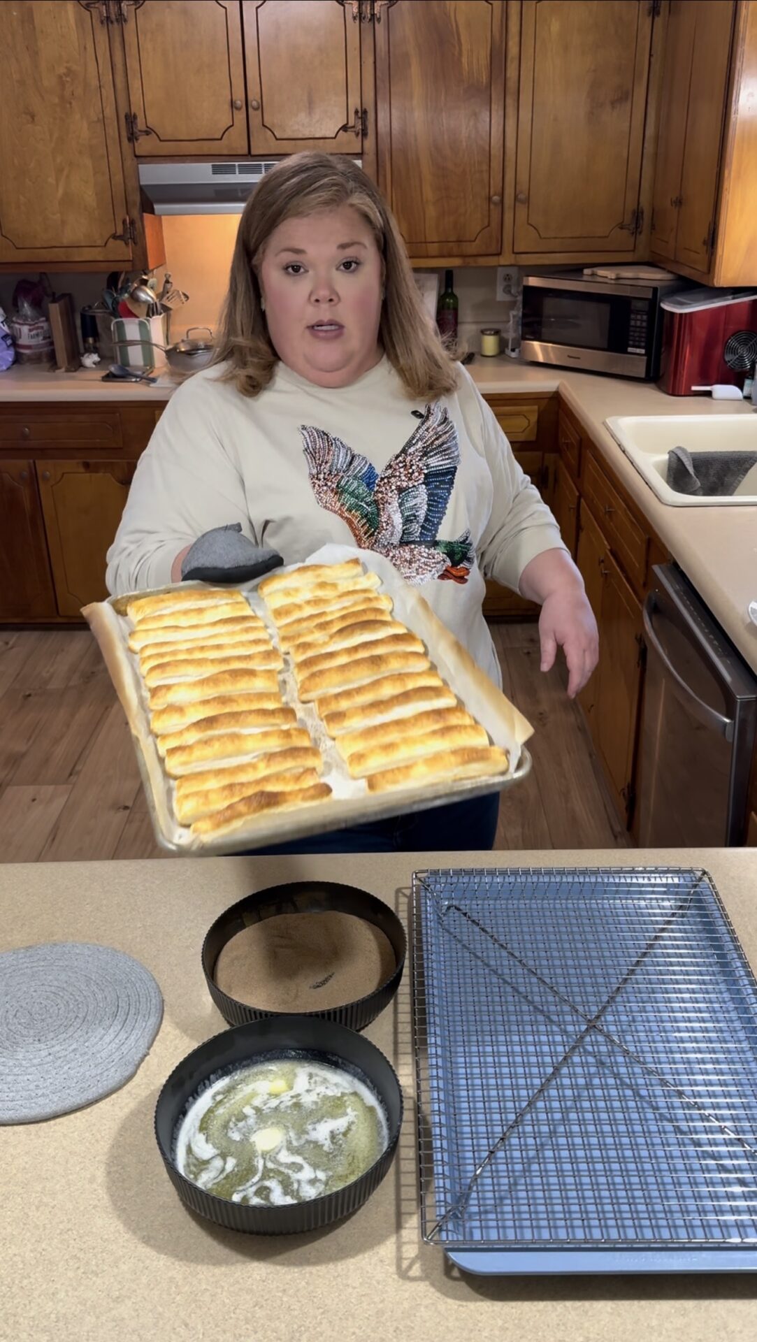 A woman in a kitchen holds a tray of freshly baked golden-brown pastries. In the foreground, there’s a cooling rack, a round pan with melted butter, and a round pan with cinnamon sugar on a countertop.
