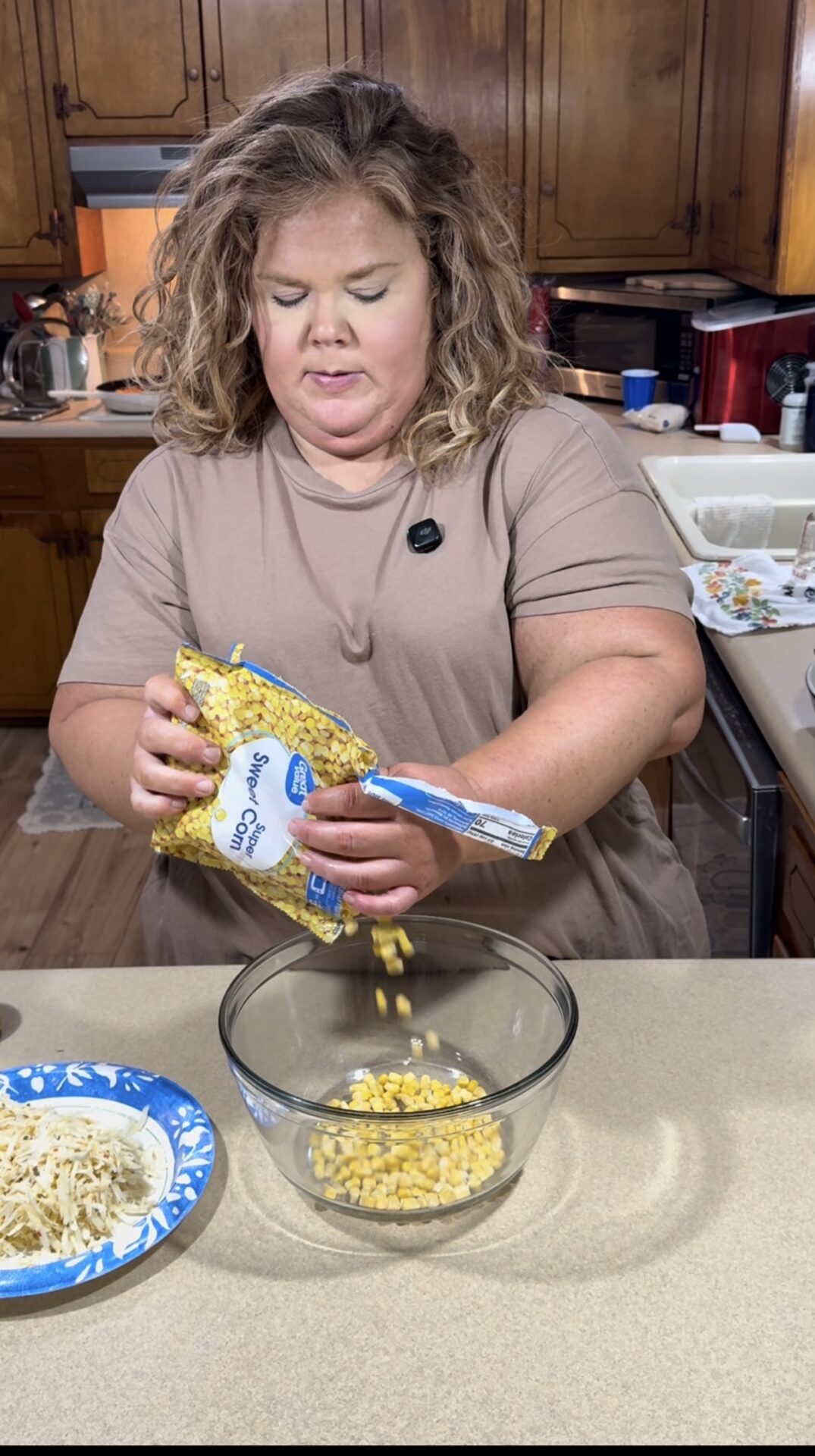 A woman with curly blonde hair in a tan shirt pours frozen corn from a bag into a glass bowl on a kitchen counter, with shredded cheese on a blue paper plate nearby. Wooden cabinets and kitchen appliances are in the background.