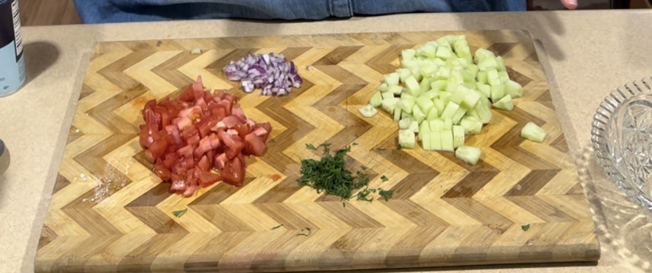 A wooden cutting board with chopped red onions, diced tomatoes, diced cucumbers, and chopped herbs arranged in small piles on a kitchen counter.