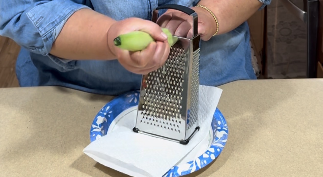 A person in a blue shirt uses a box grater to grate a peeled cucumber over a paper towel-lined plate on a kitchen counter.