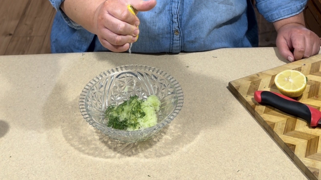 A person in a blue shirt squeezes a lemon over a glass bowl containing chopped herbs and cucumber, with a cutting board, knife, and halved lemon nearby on a countertop.