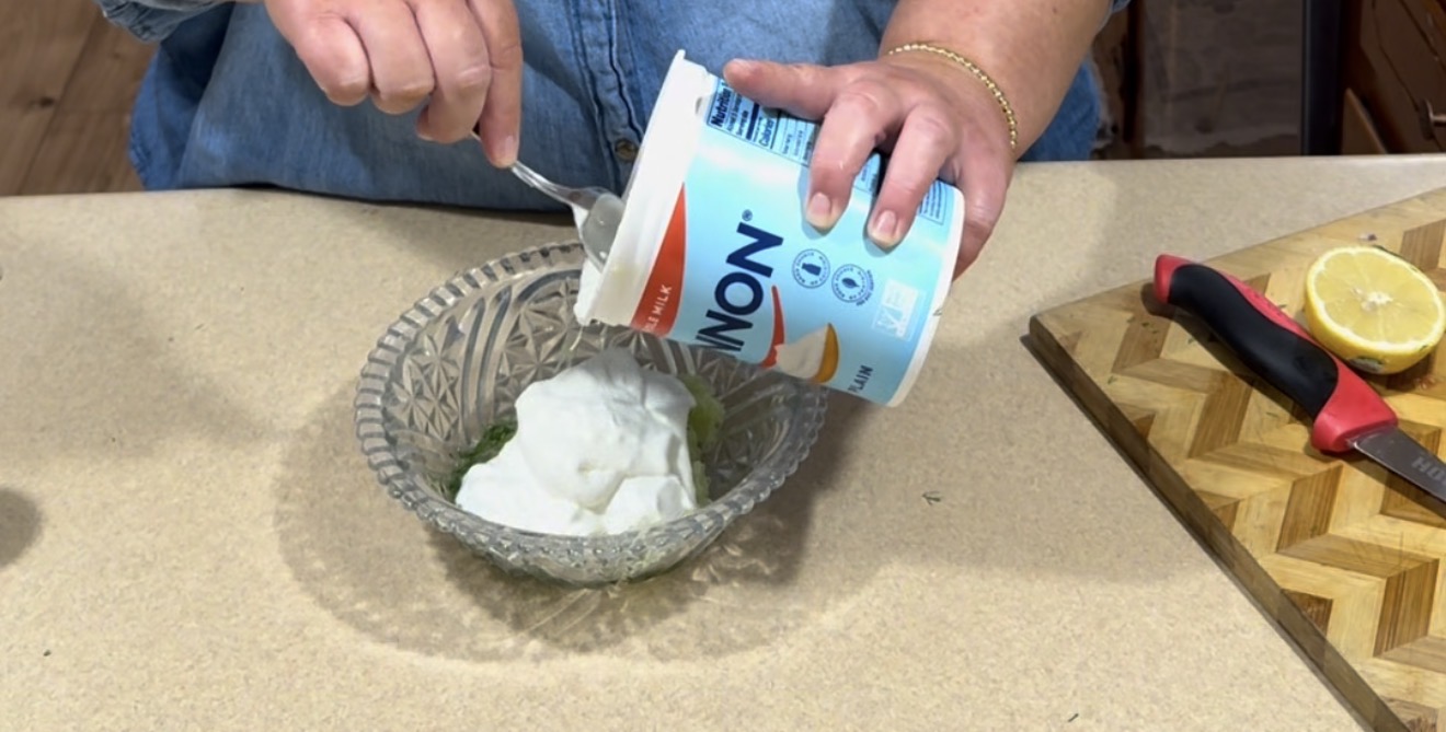 A person scoops plain non-fat yogurt from a large container into a glass bowl on a kitchen counter. Nearby, there is a cutting board with a knife and a halved lemon.