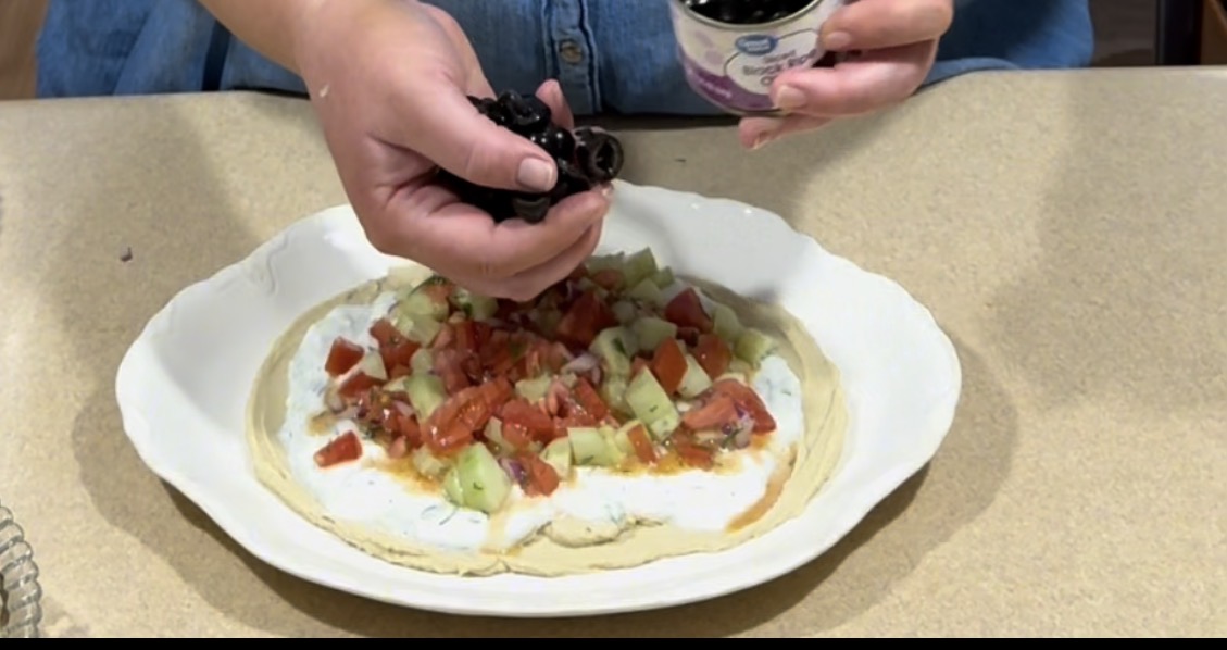A person is adding sliced black olives from a container onto a flatbread topped with a white sauce, diced tomatoes, and chopped cucumbers on a white plate.