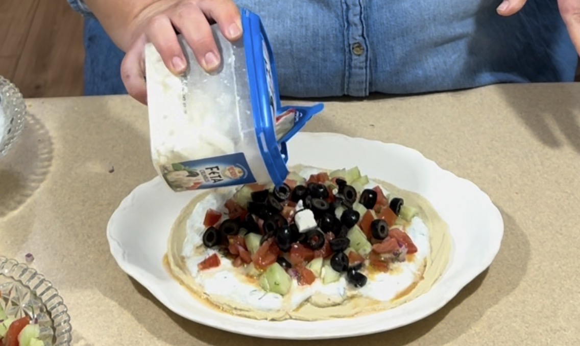 A person sprinkles crumbled feta cheese from a container onto a flatbread topped with yogurt, chopped vegetables, and black olives, on a beige kitchen counter.