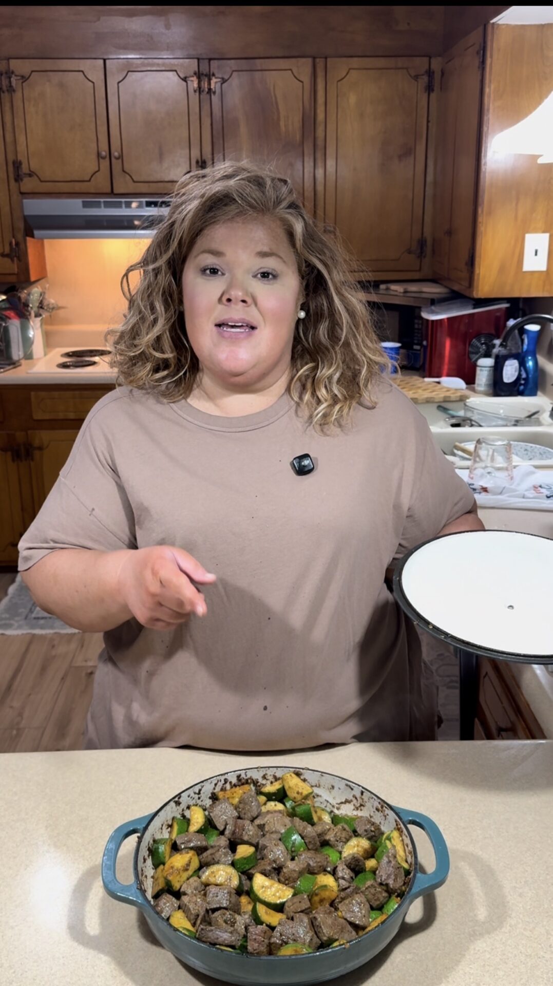 A woman with wavy hair wearing a beige shirt stands in a kitchen, holding a white plate and pointing at a pan of cooked meat and vegetables on the counter in front of her. The kitchen has wooden cabinets and a stove.