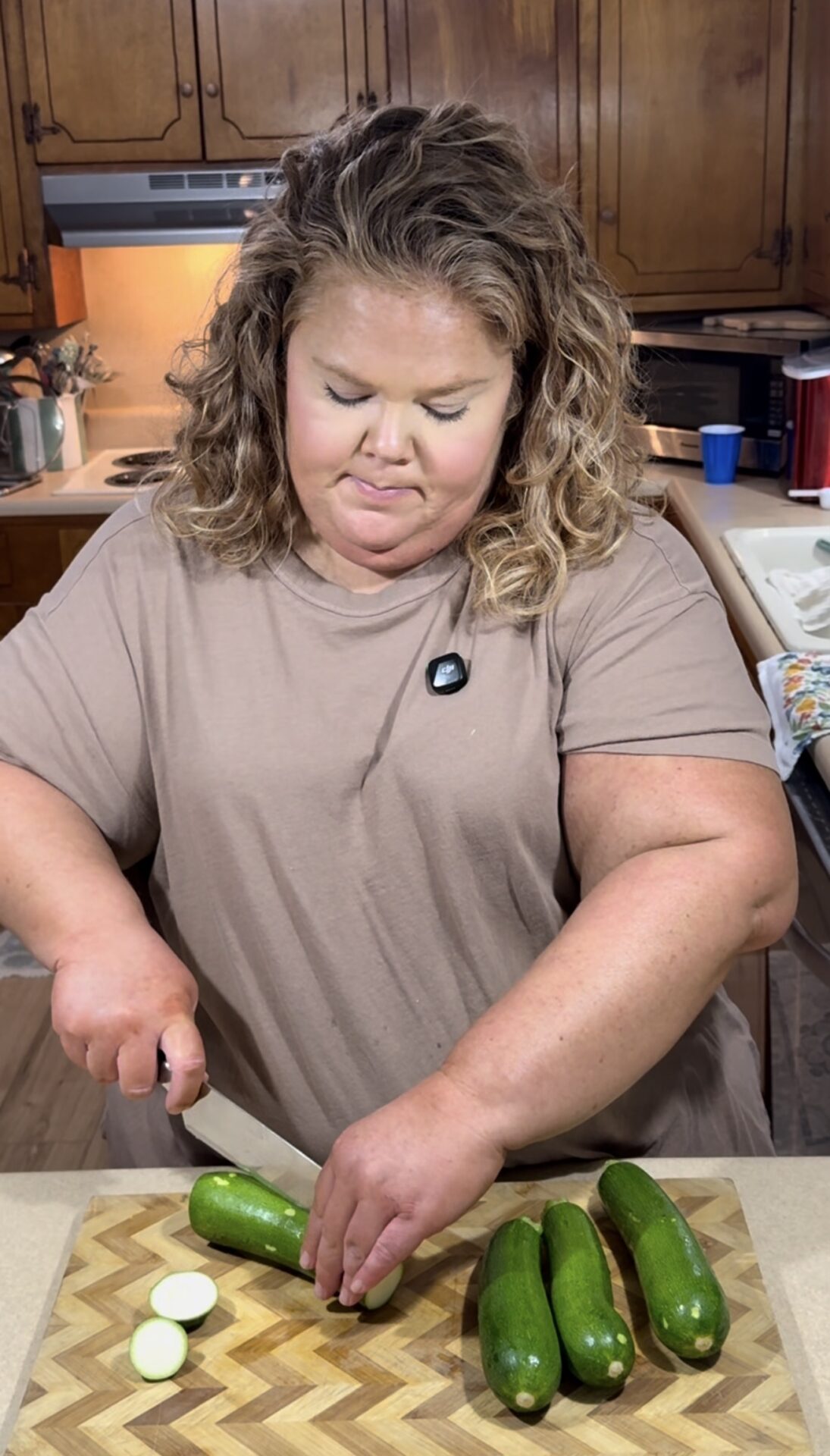 A woman with curly hair, wearing a beige shirt, slices zucchini on a cutting board in a kitchen with wooden cabinets. She is focused on her task and standing at a counter.