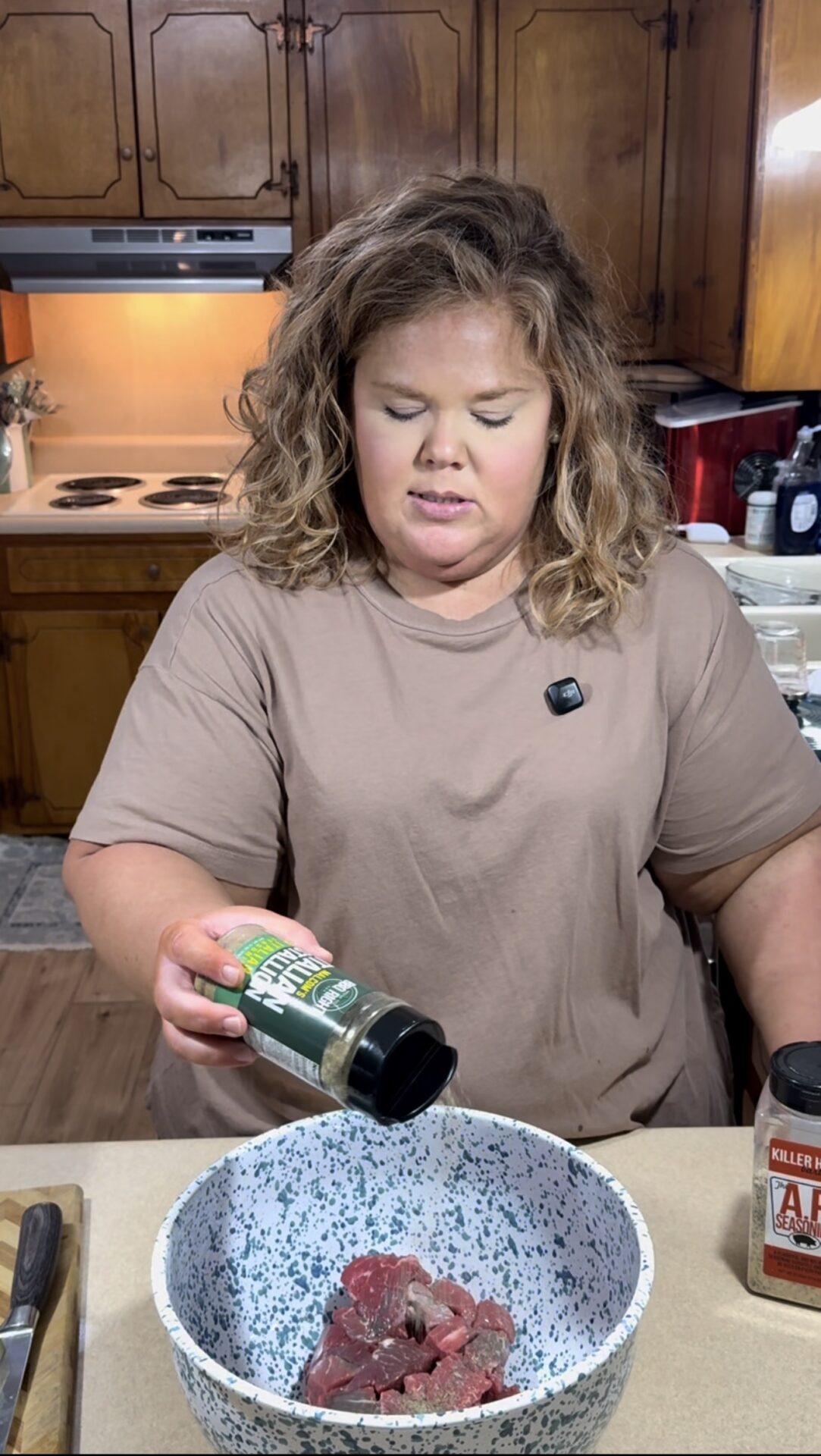 A woman in a tan shirt sprinkles seasoning from a green-labeled container onto raw meat in a white and blue speckled bowl in a kitchen with wooden cabinets. A spice jar and knife are visible on the counter.
