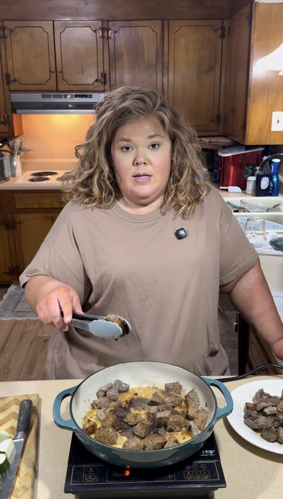 A woman with curly hair and a tan shirt cooks beef pieces in a pan on a stovetop, holding tongs. She stands in a home kitchen with wooden cabinets and various kitchen items in the background.
