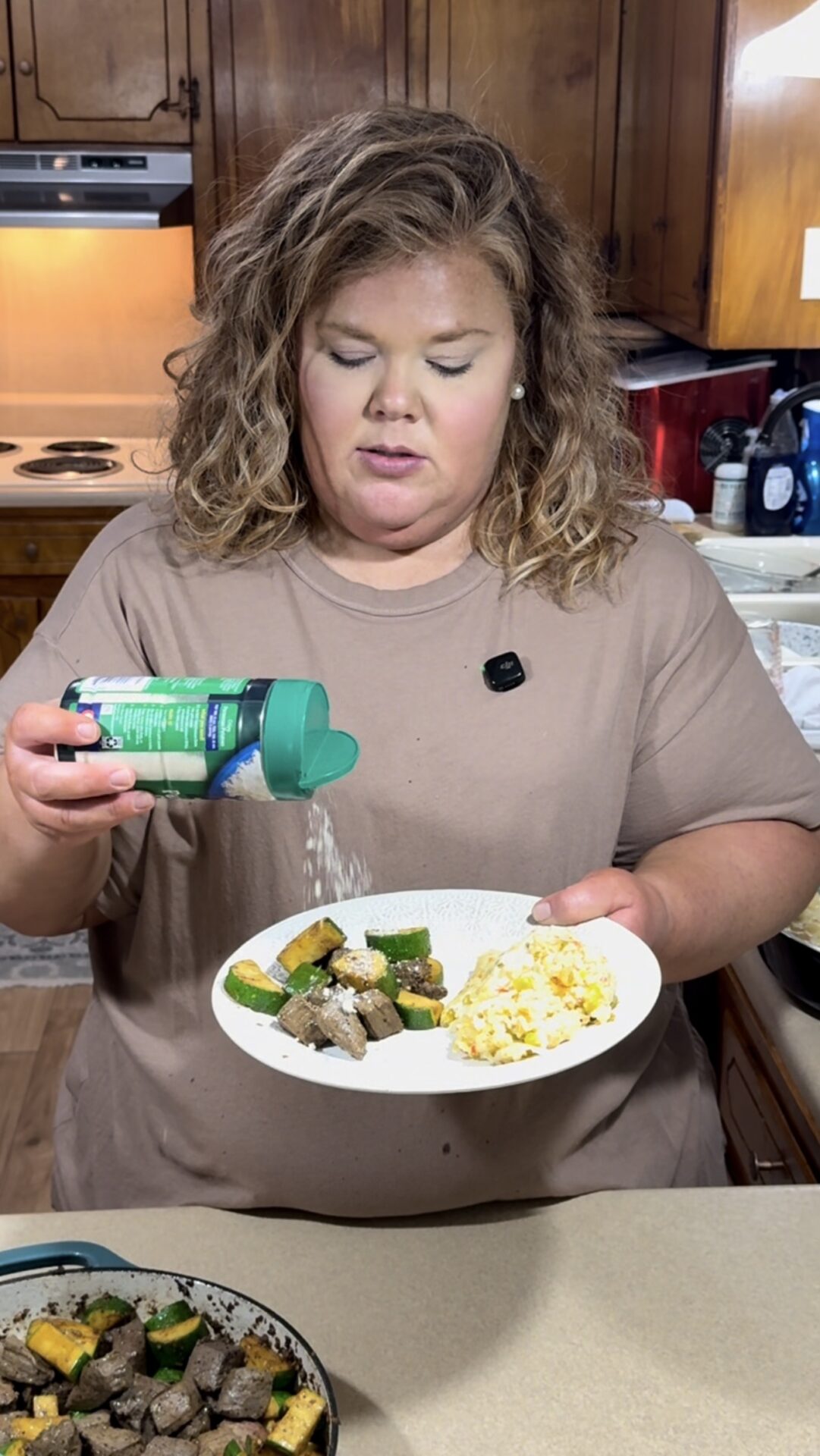 A woman with wavy hair and a tan shirt sprinkles grated cheese onto a plate of scrambled eggs, steak, and sautéed vegetables in a kitchen with wooden cabinets.