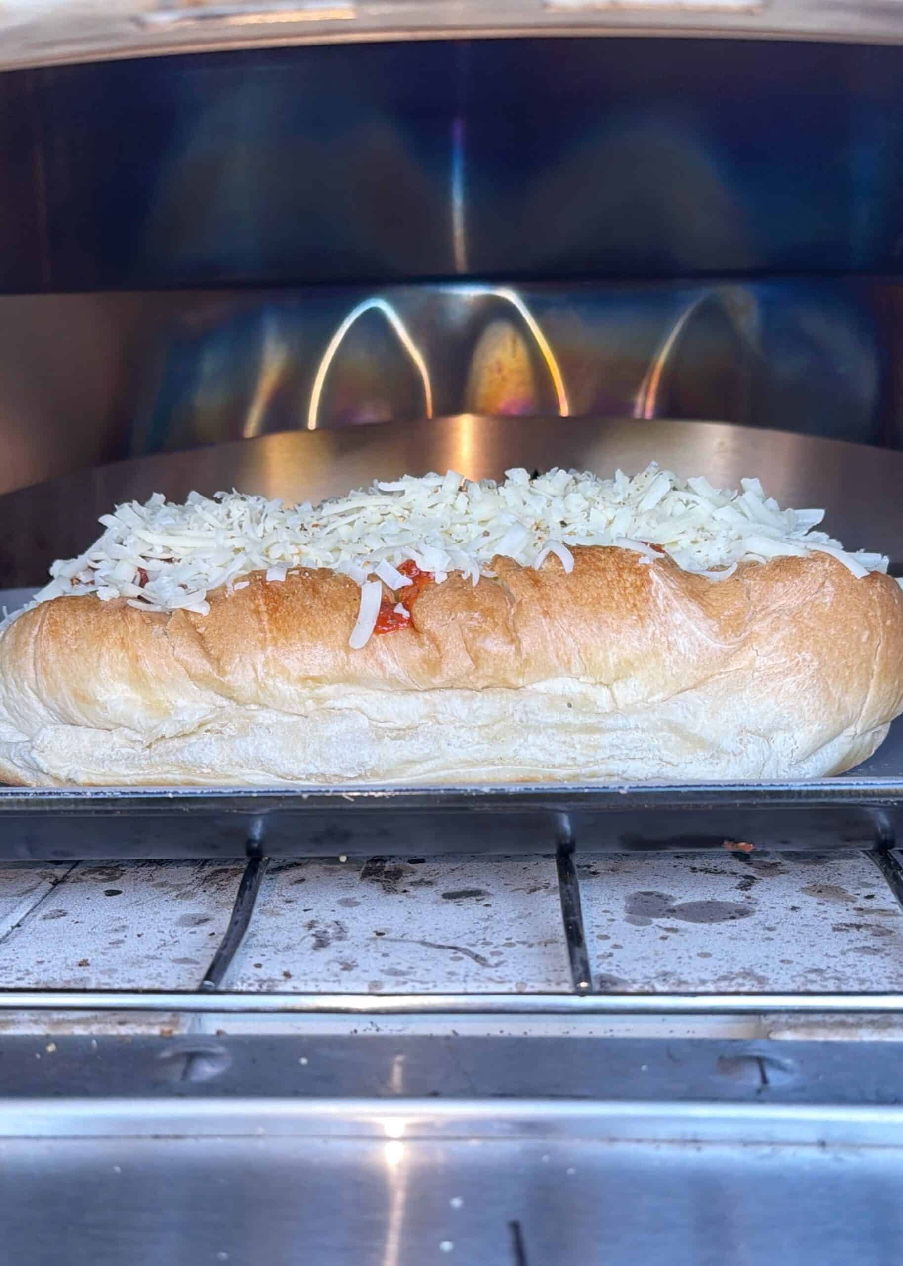 A sandwich roll topped with shredded cheese is being toasted in an oven, with the cheese beginning to melt.