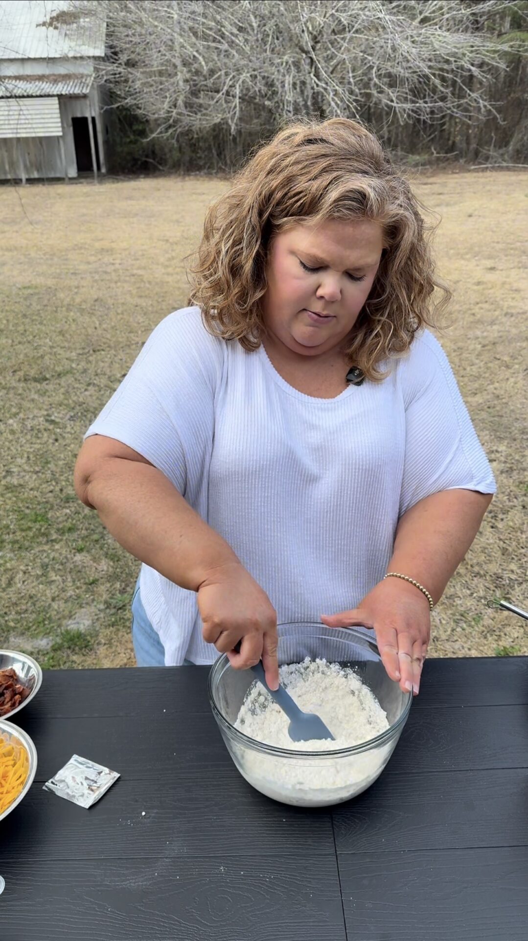A woman with curly hair in a white shirt mixes flour in a glass bowl on an outdoor table, with bowls of ingredients nearby and a grassy yard with trees in the background.
