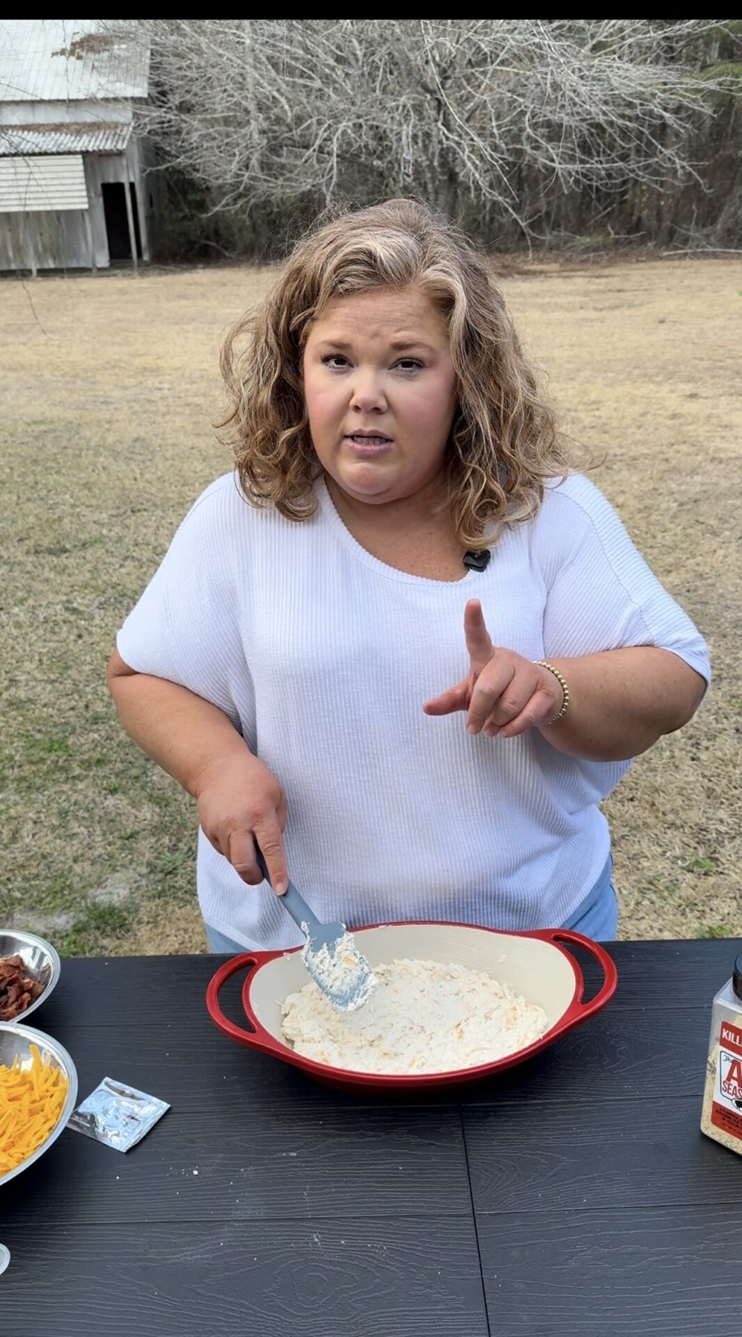 A woman with curly hair in a white shirt mixes ingredients in a red baking dish outdoors. She looks at the camera, raising one finger, with bowls of cheese and bacon and a spice jar on the black table in front of her.