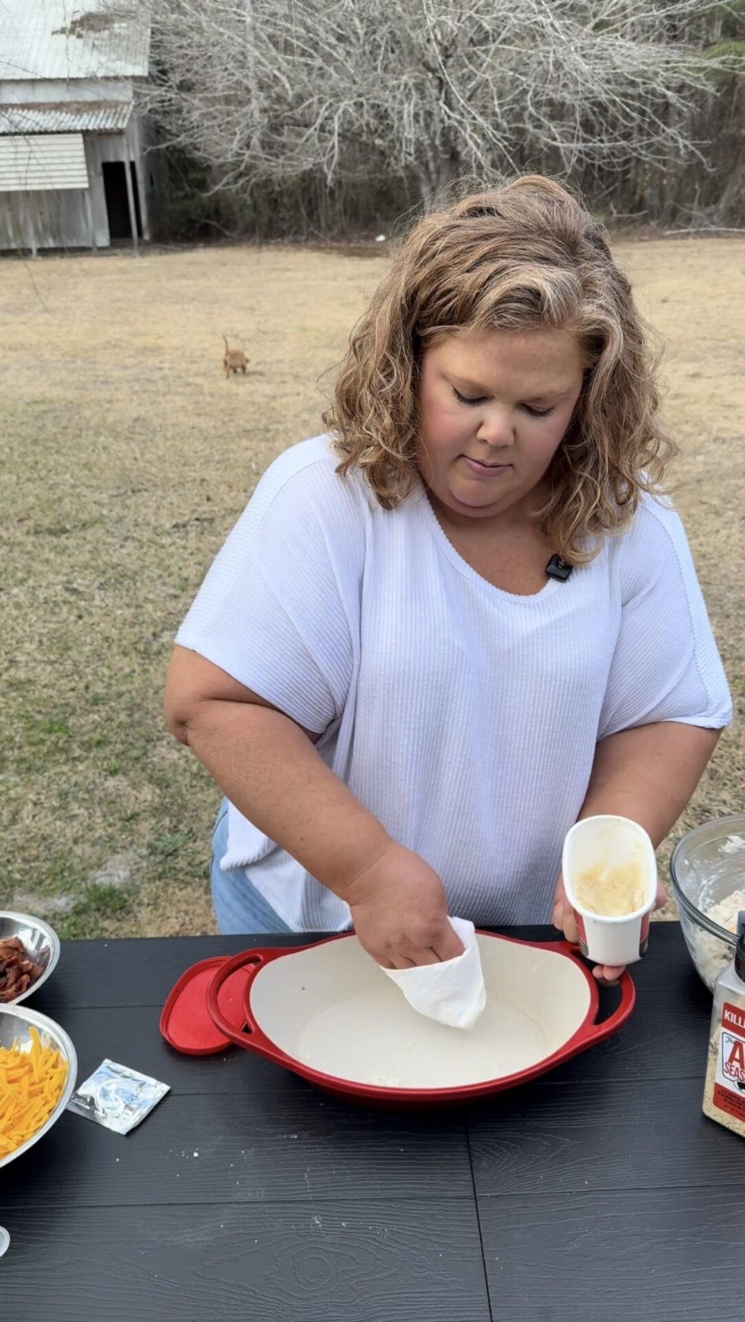 A woman with curly hair in a white shirt greases a red baking dish outdoors on a black table surrounded by bowls of cheese, bacon, and other ingredients. Leafless trees and a barn are in the background.