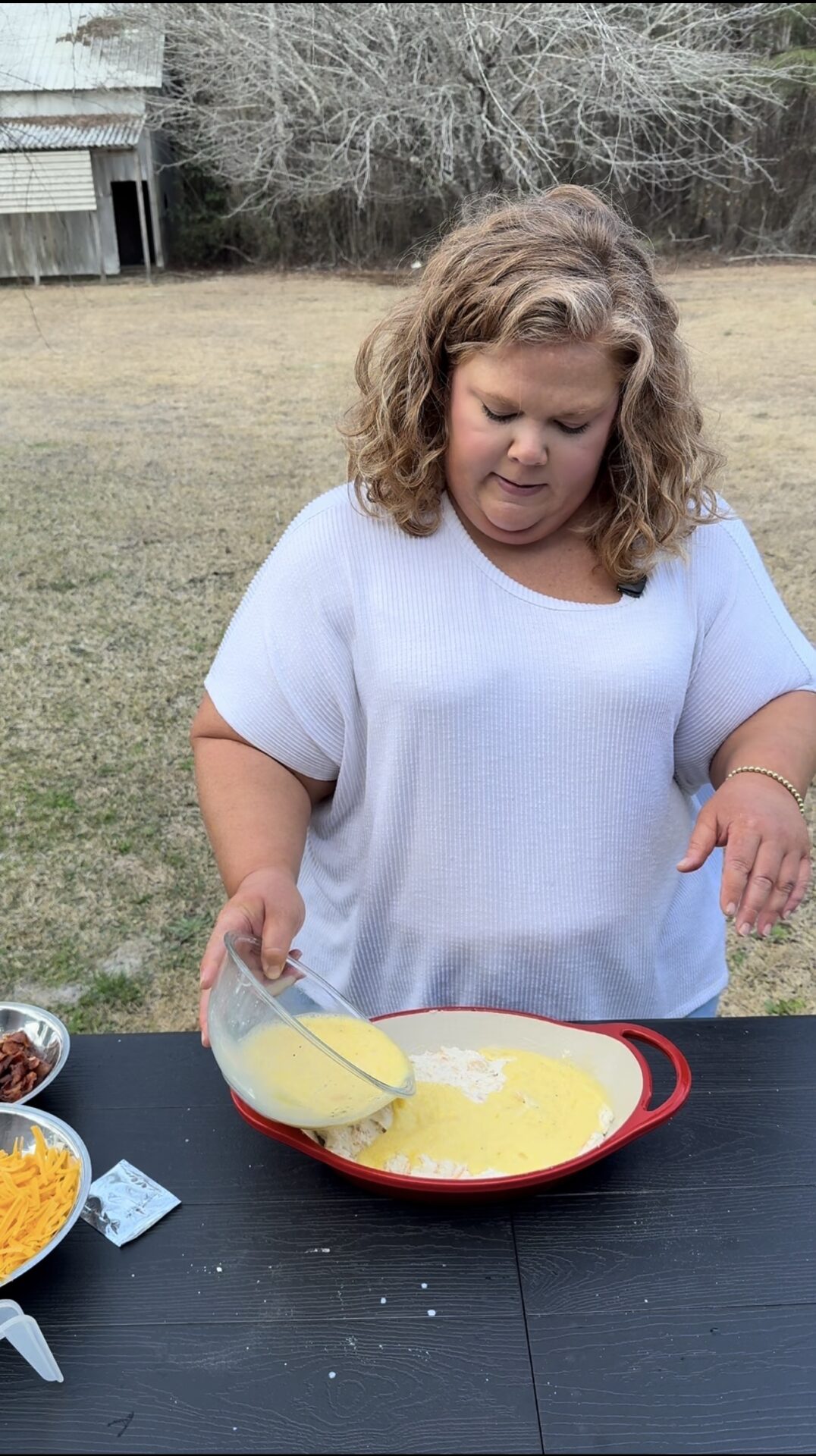 A woman with curly hair wearing a white shirt pours a yellow batter or mixture from a glass bowl into a red dish on a black table outdoors. Bowls of cheese, bacon, and other ingredients are nearby.