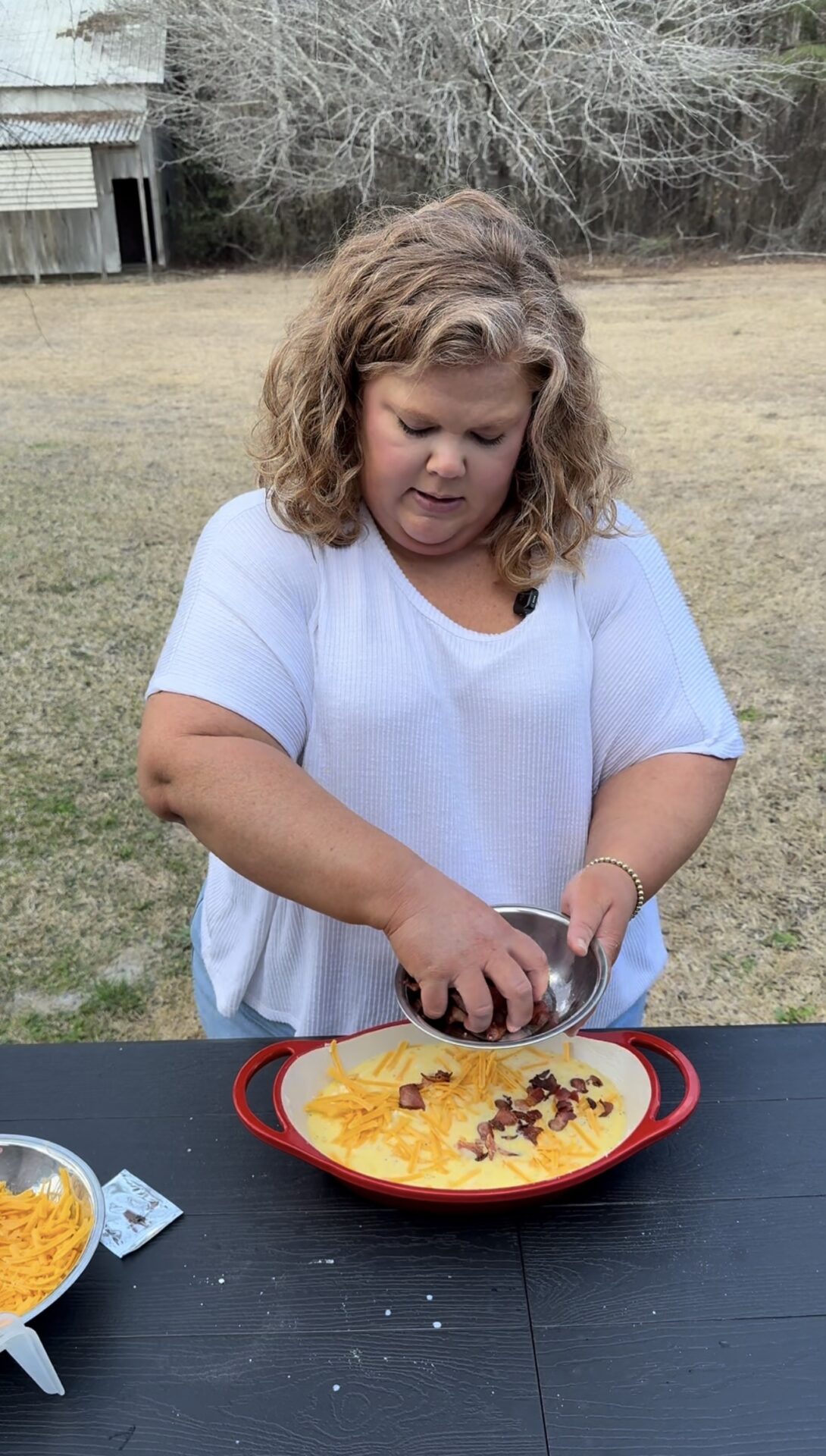A woman with curly hair in a white shirt stands outside, sprinkling toppings from a bowl onto a casserole dish filled with cheese, on a black table. A grassy yard and trees are visible in the background.