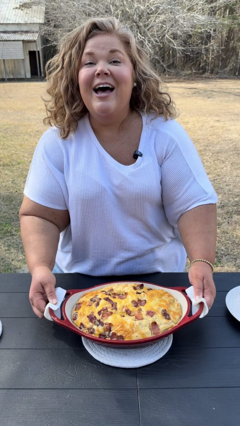 A smiling woman with curly hair holds a red dish filled with a cheesy baked casserole, standing at an outdoor table with grass and trees in the background.
