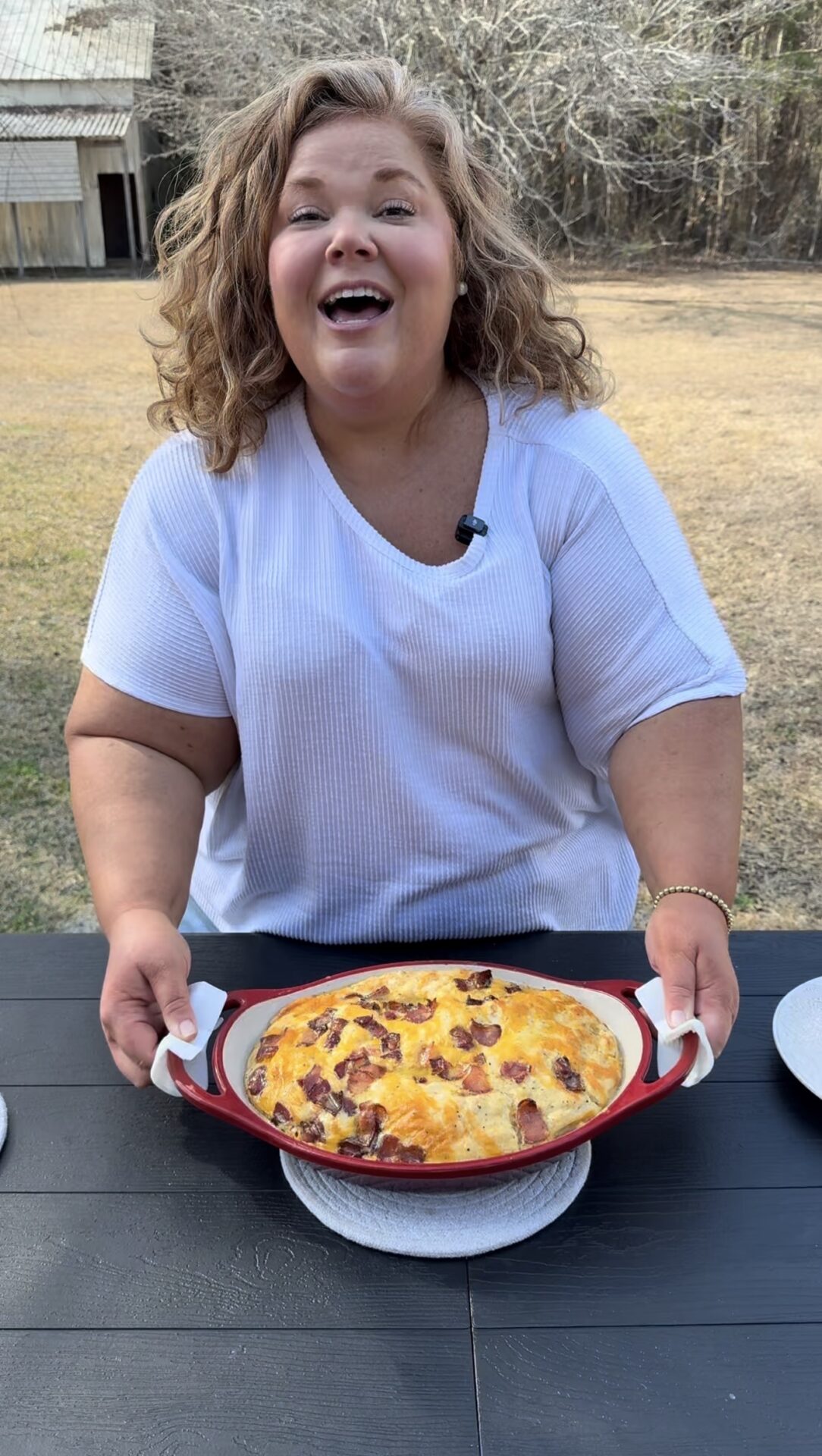 A smiling woman with curly hair holds a red dish filled with a cheesy baked casserole, standing at an outdoor table with grass and trees in the background.
