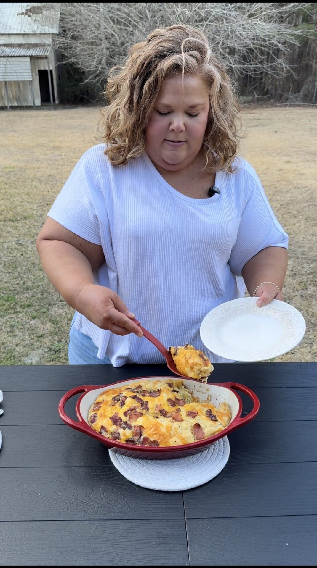 A woman in a white shirt serves a cheesy baked casserole onto a plate using a red spoon. She is standing outdoors by a black table, with grass and trees visible in the background.