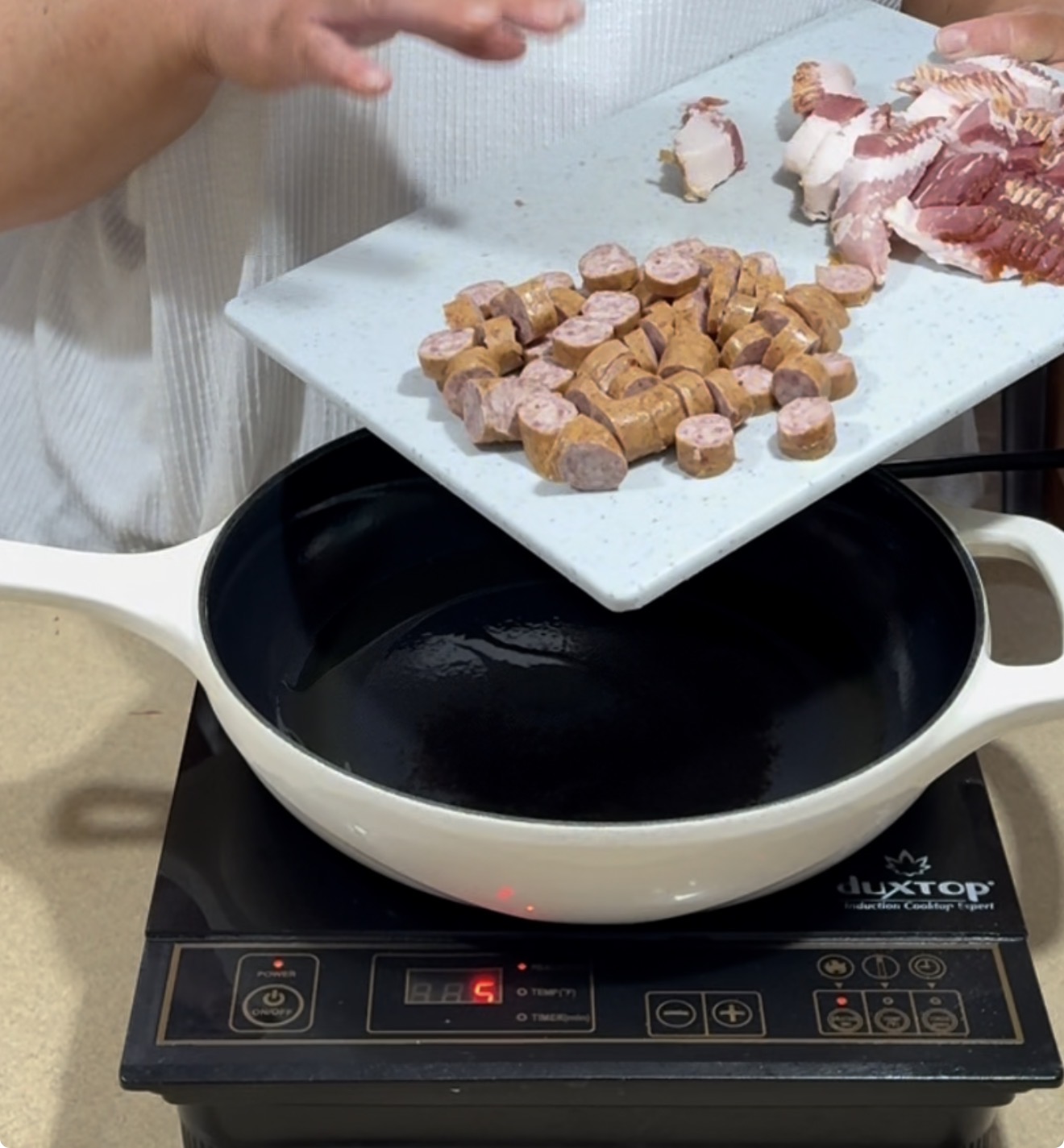 A person prepares to add sliced sausage and bacon from a cutting board into a white pan on an electric induction cooktop.