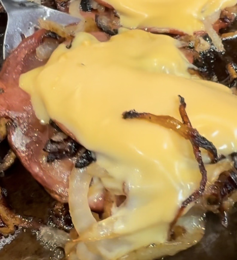 Slices of fried bologna topped with grilled onions and melted yellow cheese, cooking on a skillet. The cheese is slightly melted over the onions and bologna, giving a glossy appearance.
