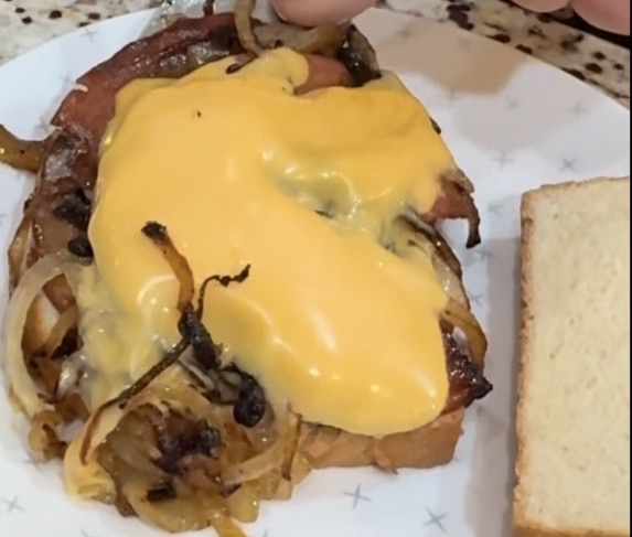 An open-faced sandwich on white bread topped with grilled onions, cooked meat, and melted yellow cheese sits on a white plate with a second slice of plain bread beside it.