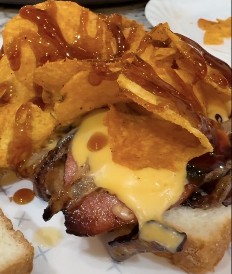 A close-up of a sandwich with melted cheese, bacon, potato chips, and barbecue sauce on bread, served on a white plate.