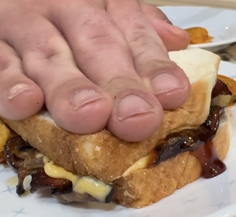 A close-up of a persons hand pressing down on a sandwich filled with bacon, melted cheese, and sauce, on a white plate.