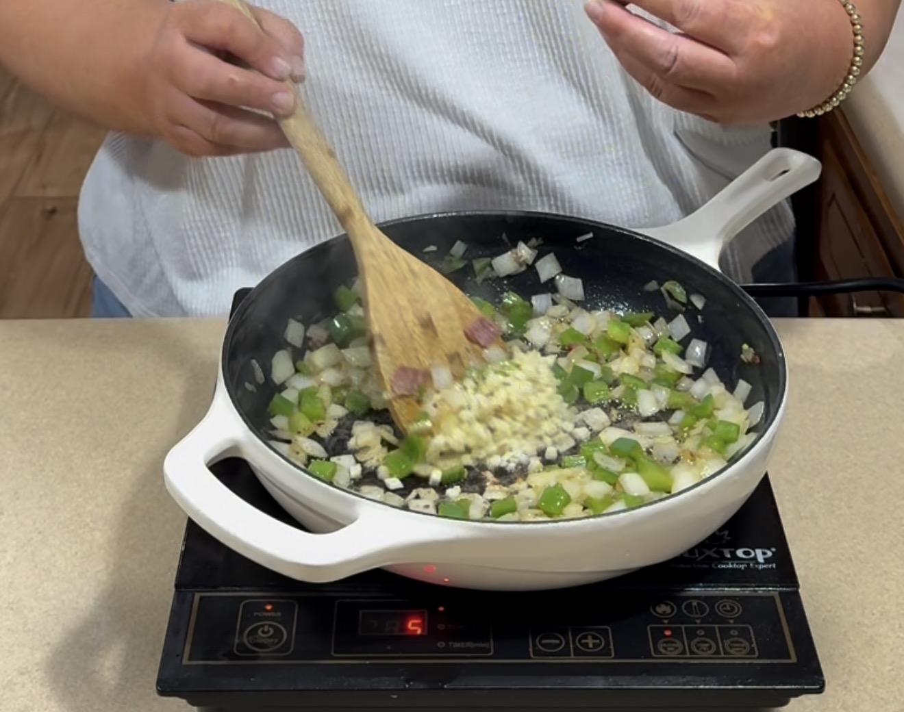 A person cooks diced onions, green peppers, and garlic in a white skillet on an electric stovetop, stirring with a wooden spatula.