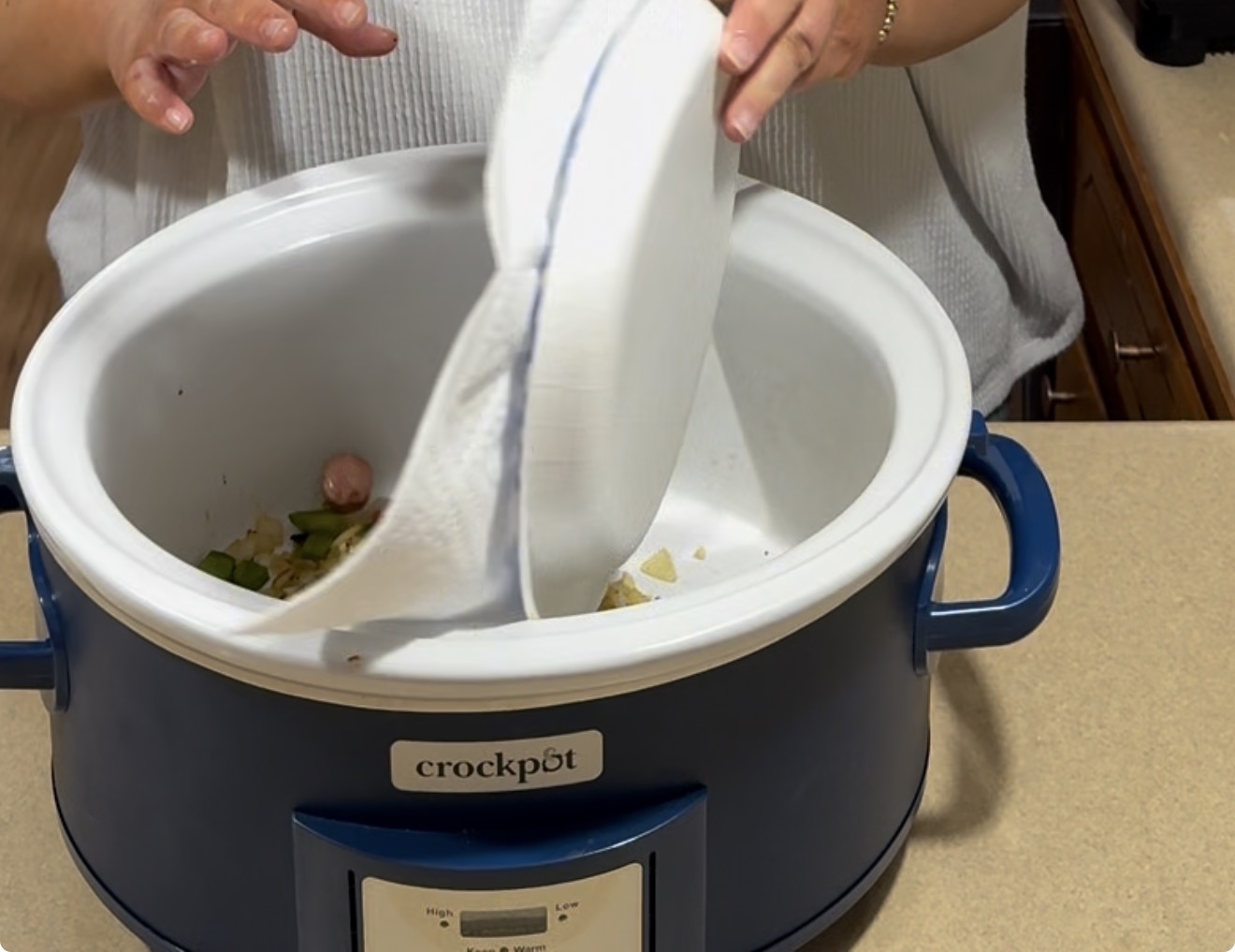 A person pours chopped vegetables from a white cutting board into a blue Crockpot slow cooker on a kitchen counter.