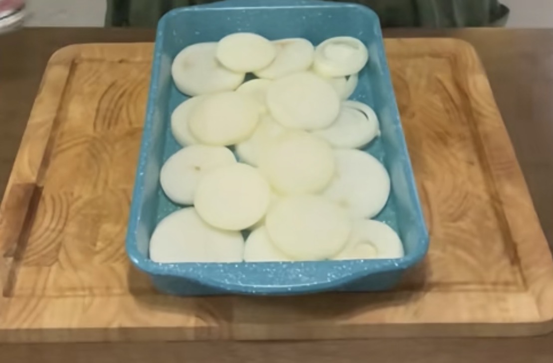A turquoise baking dish filled with raw, sliced onions sits on a wooden cutting board.
