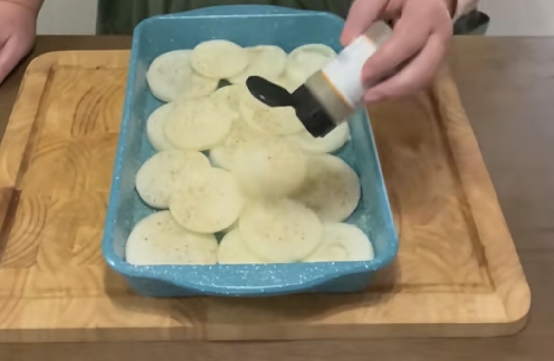 A person sprinkles seasoning from a container onto a layer of sliced potatoes arranged in a blue baking dish on a wooden cutting board.