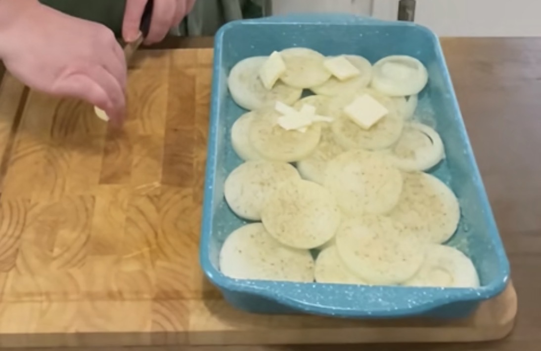 A person slices butter on a wooden cutting board next to a blue baking dish filled with sliced onions, some topped with butter pats and seasoning.