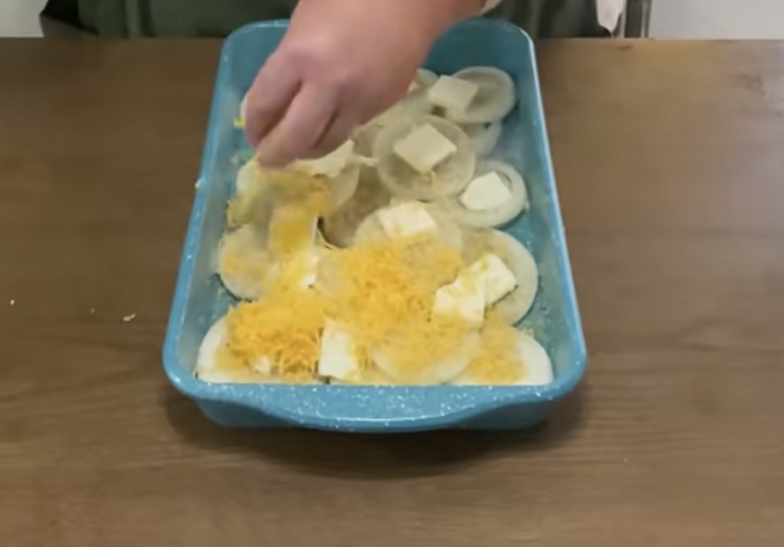 A hand sprinkles shredded cheese over sliced potatoes and butter in a blue baking dish, preparing a layered potato casserole on a wooden table.