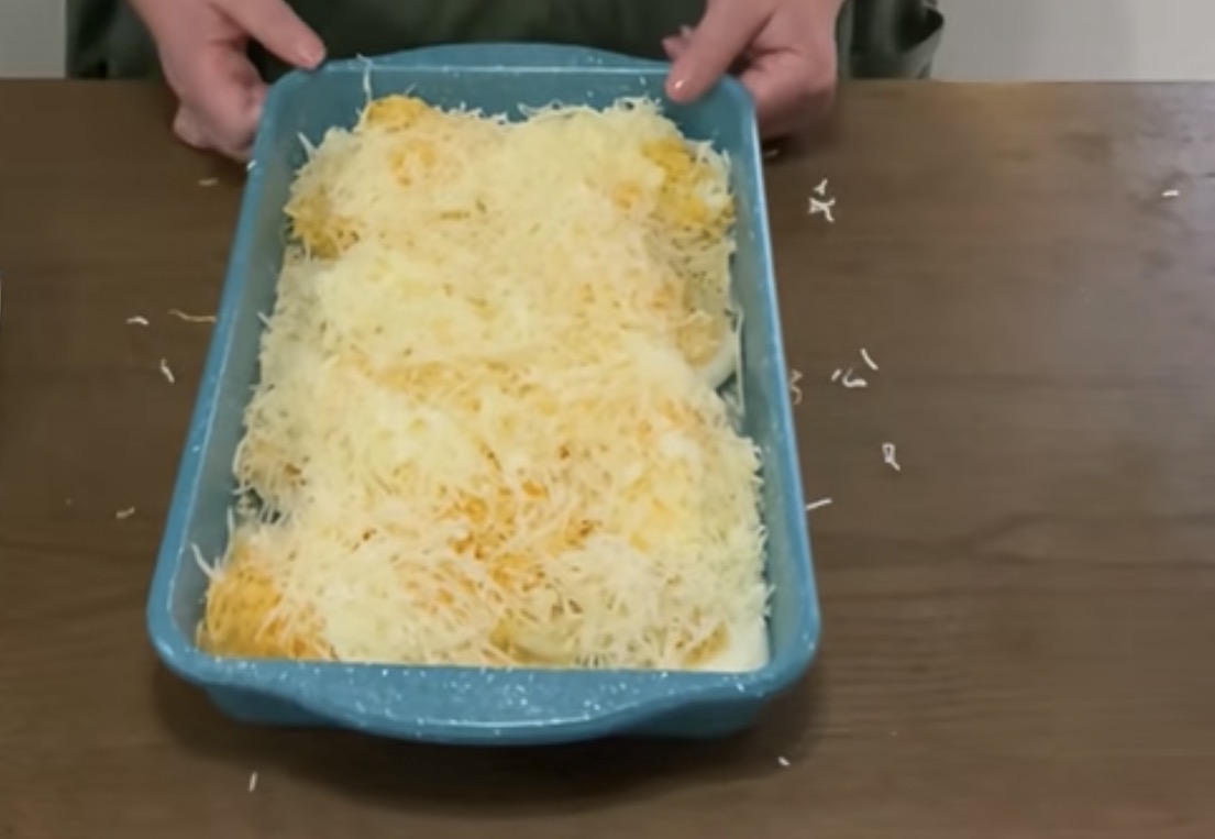 A person holds a blue baking dish filled with sliced potatoes topped with a thick layer of grated cheese, ready to be baked. Some cheese is scattered on the wooden table.
