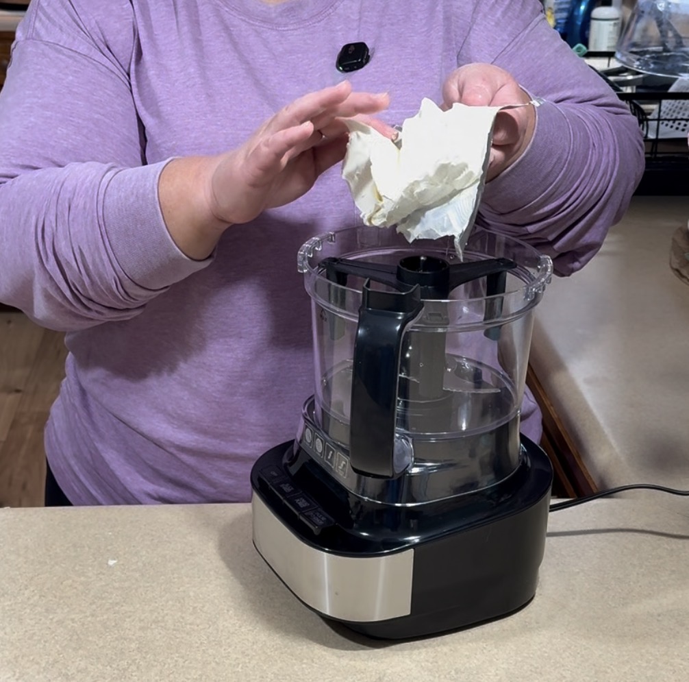 A person in a purple shirt adds a large chunk of cream cheese to a food processor on a kitchen counter.