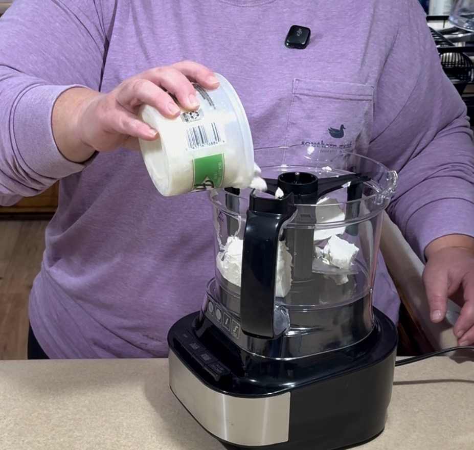 A person in a purple shirt is pouring a container of cream cheese into a food processor on a kitchen counter.