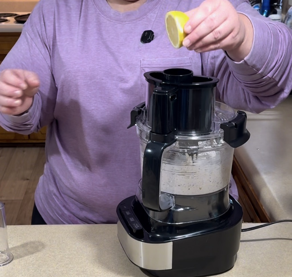 A person in a purple top squeezes a lemon half over a food processor on a kitchen counter, preparing an ingredient mixture.