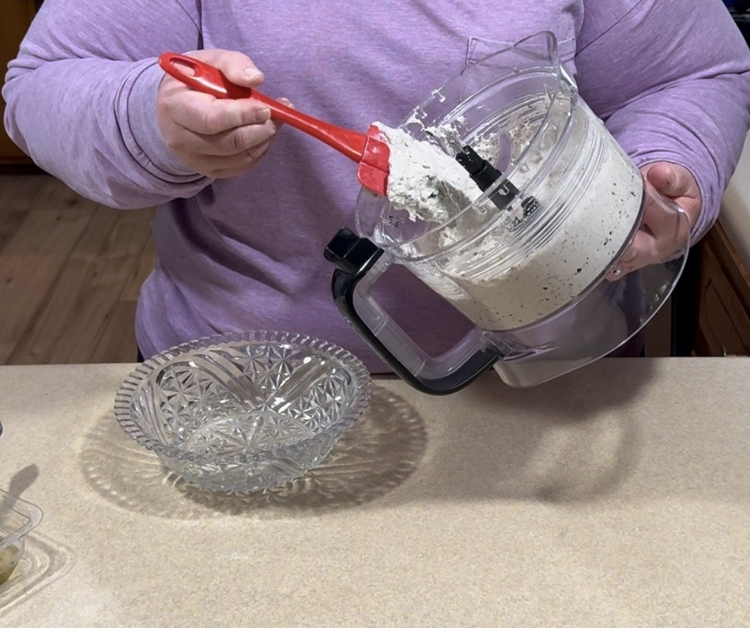 A person in a purple shirt uses a red spatula to scrape a thick, white mixture with specks from a food processor bowl into a clear, patterned glass serving bowl on a kitchen counter.