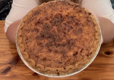 A person holding a large, round pie in a metal pie tin above a wooden table; the pie has a golden-brown, slightly uneven, crumbly crust.