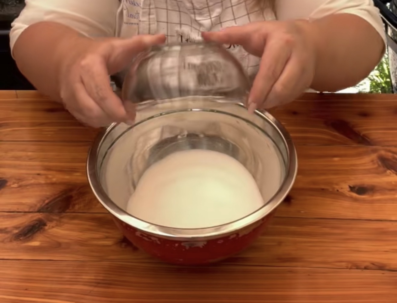 A person holding a clear glass bowl over a larger mixing bowl filled with white powder, possibly sugar or flour, on a wooden table.