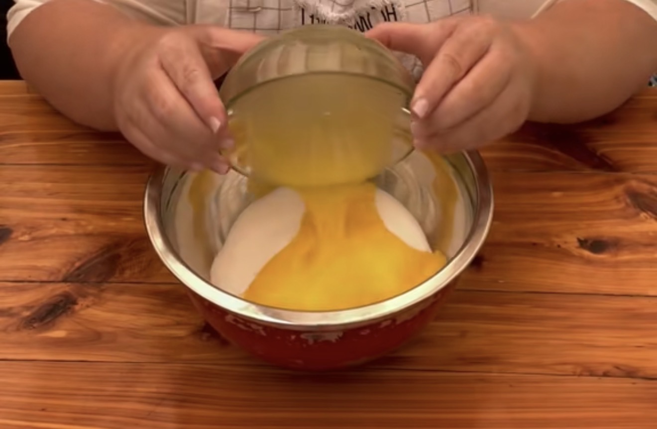 A person pours a bowl of egg yolks into a larger bowl filled with sugar on a wooden table, preparing ingredients for baking.