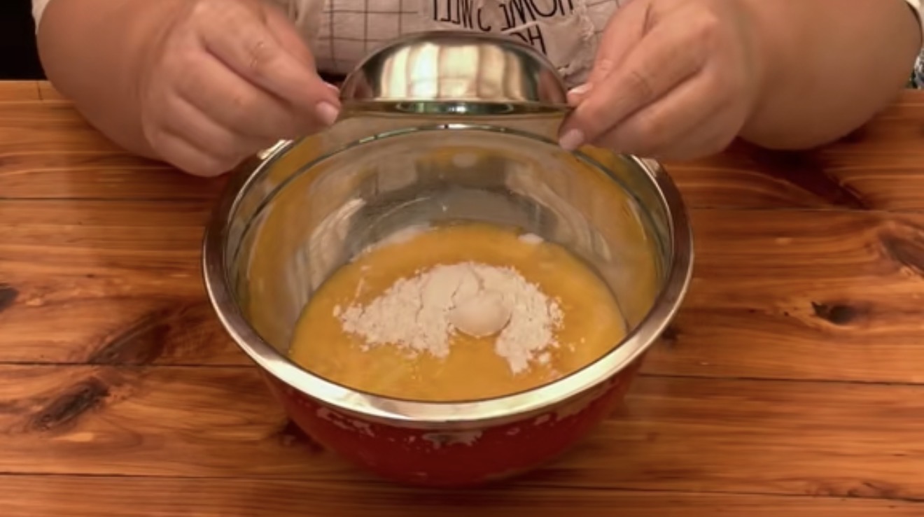 A person holds a metal bowl over a larger mixing bowl containing flour and beaten eggs, preparing ingredients on a wooden table.