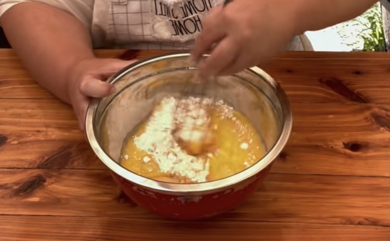 A person mixes flour and eggs in a red bowl with a whisk on a wooden table.