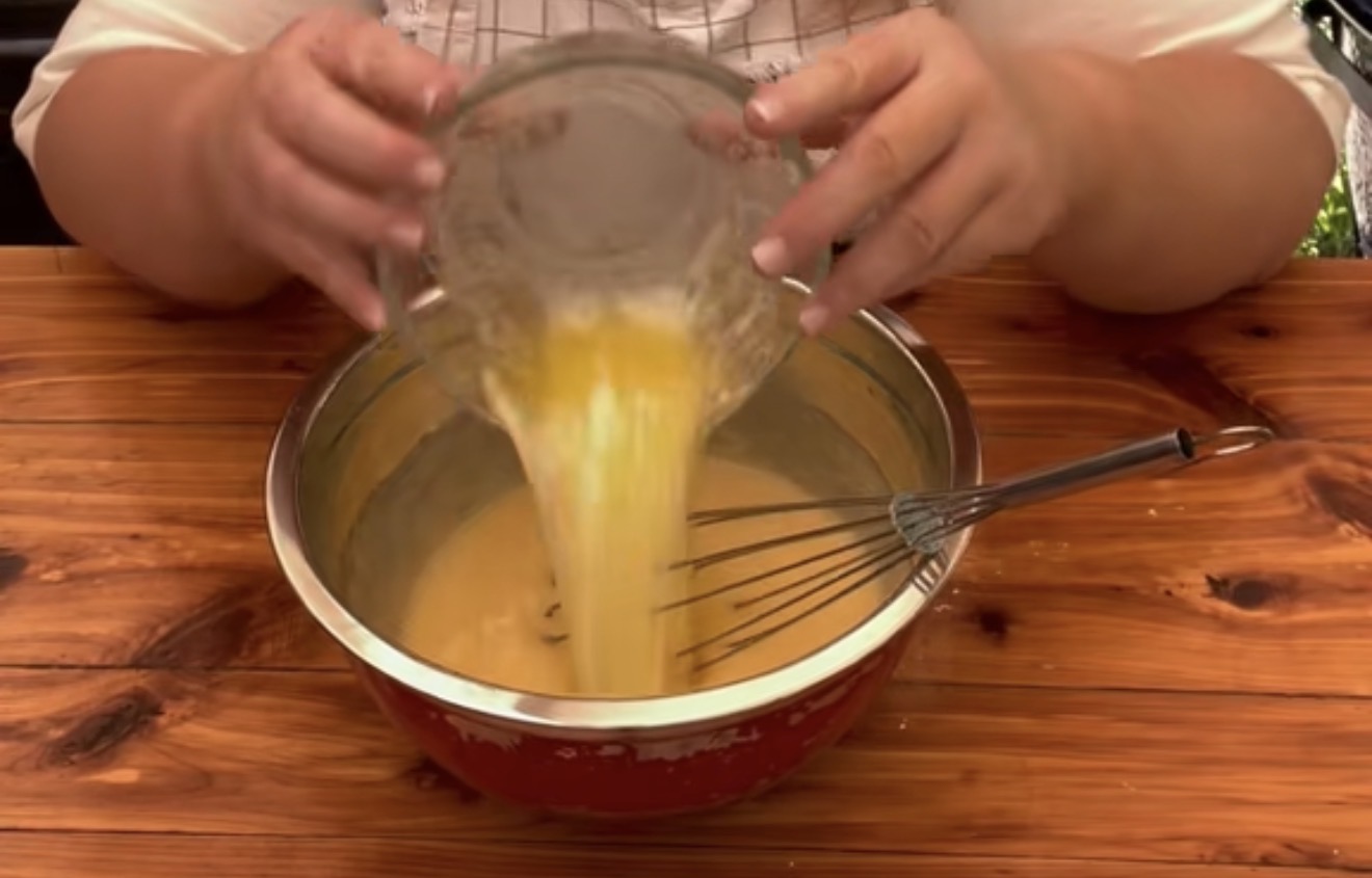 A person pours melted butter from a glass bowl into a red mixing bowl with batter and a whisk, on a wooden table.