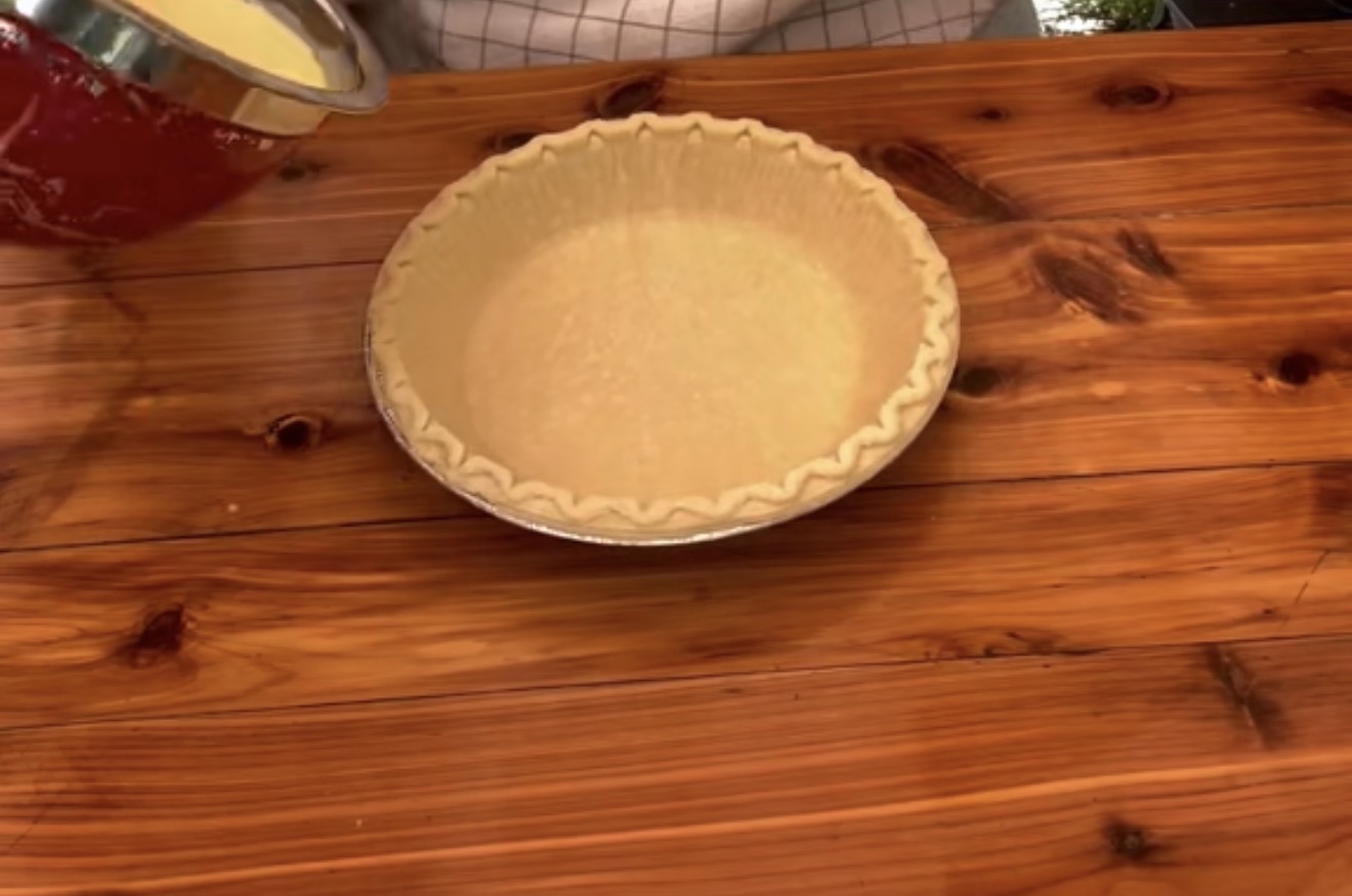 A person is about to pour a creamy mixture from a bowl into an unbaked pie crust that sits on a rustic wooden table.