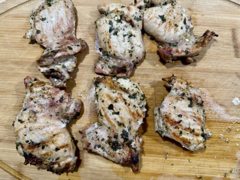 Six pieces of grilled, marinated chicken thighs with visible herbs are arranged on a wooden cutting board. The chicken has a slightly charred, golden-brown appearance.
