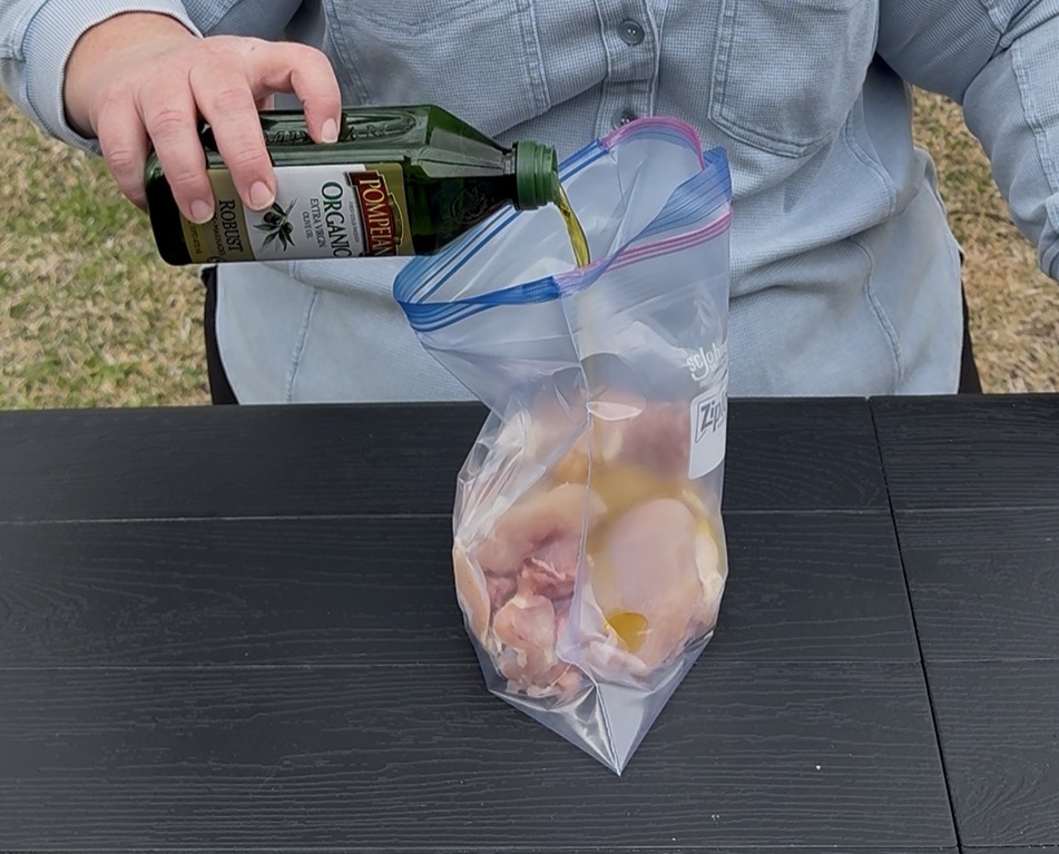 A person pours olive oil from a bottle into a resealable plastic bag filled with raw chicken pieces on a black table.
